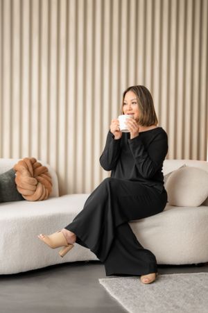 Someone in a black dress relaxes on a white couch while sipping from a coffee cup in a modern living room setting.