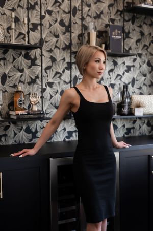 Woman in elegant black dress standing at a modern bar with stylized floral wallpaper in background.
