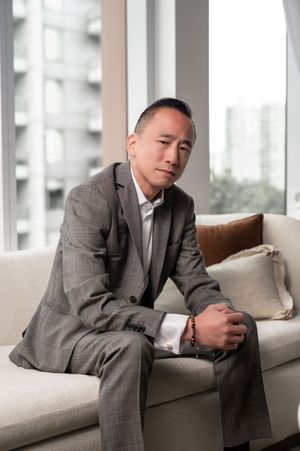 Professional wearing a gray plaid suit sits casually on a modern beige couch near large windows in an urban setting.