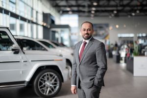 Business professional in grey suit standing next to luxury vehicle in modern showroom.