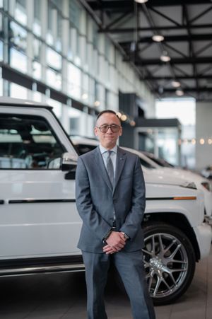 Businessman in grey suit and glasses standing beside white luxury SUV in dealership.