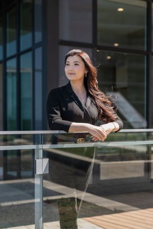 Professional woman in dark business attire leans on glass railing outside modern office building on sunny day.