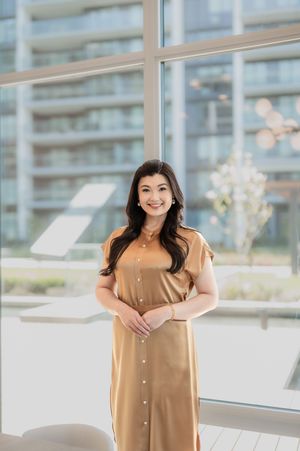 Person in a camel-colored button-down dress stands in a bright, modern room with city views through large windows.