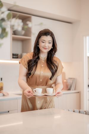 A person in a beige dress serves coffee cups on a tray in a modern, minimalist kitchen setting.