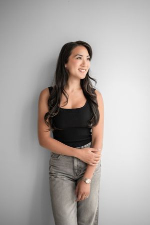 A figure in a black tank top and light wash jeans poses casually against a white wall in a minimalist setting.