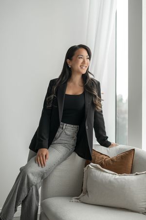Professional woman in black blazer and gray pants poses elegantly on white couch with brown accent pillows.