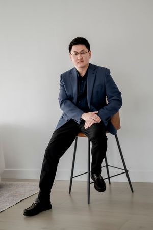 Professional portrait of a person in a navy blazer seated on a modern black stool against a light gray wall.