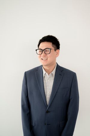 Professional smiling person wearing glasses, dressed in a navy suit and patterned shirt against a light background.