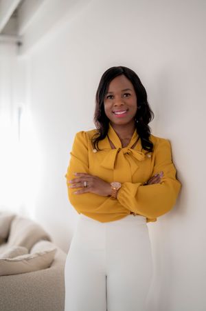 Professional woman in a mustard yellow bow blouse and white pants poses confidently with arms crossed against white wall.