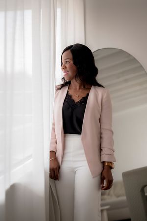 Professional portrait with stylish light pink blazer, black lace top and white pants against bright window light.