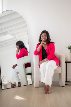 Professional photo showing a person wearing a bright pink blazer and white pants seated in front of a large mirror in a minimalist room.