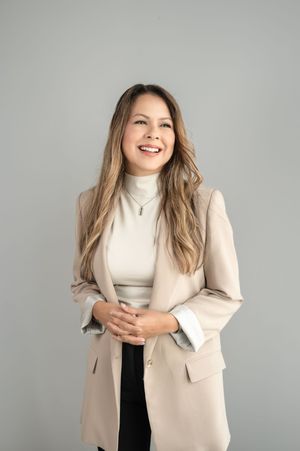 Professional portrait of someone in a beige blazer and white turtleneck smiling against a gray background.