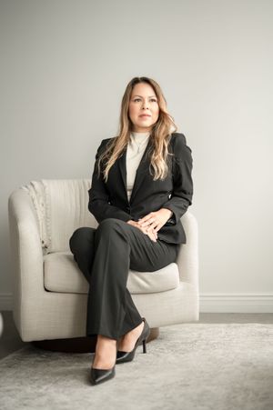 Professional in a black business suit sits confidently in a light gray upholstered armchair against neutral wall.