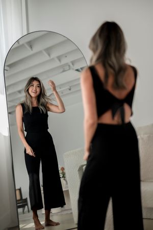 Someone wearing an elegant black jumpsuit with cutout back detail poses in front of a round mirror in a bright room.