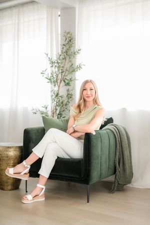 A person sits in a modern green velvet armchair with white curtains and potted plant in a bright minimalist room.