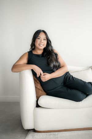 Woman in black dress poses elegantly on white chaise lounge sofa against neutral background.