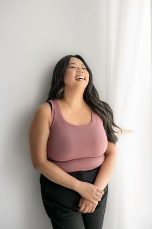 A person wearing a mauve tank top and black pants poses against a white wall with joyful expressions.