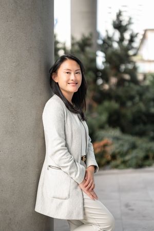Professional portrait of someone in a light gray blazer and white pants posing against concrete columns outdoors.