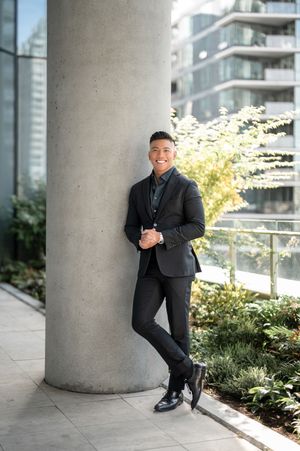 Professional in a dark suit leans against a concrete pillar with modern architecture and greenery in the background.