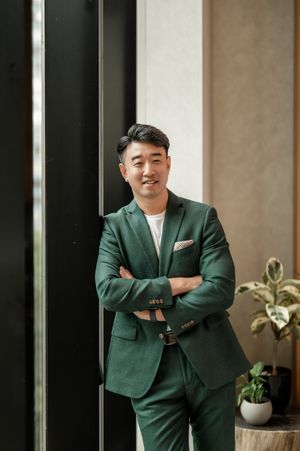 Professional in a dark green business suit poses confidently against a modern interior with windows and plants.