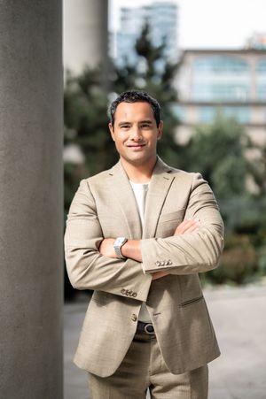 Professional portrait of a person in a beige suit jacket standing with crossed arms against a blurred outdoor background.