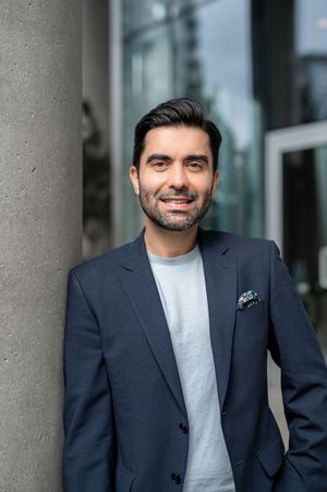 Professional portrait of someone in a navy blazer and light gray t-shirt standing against a concrete pillar outdoors.