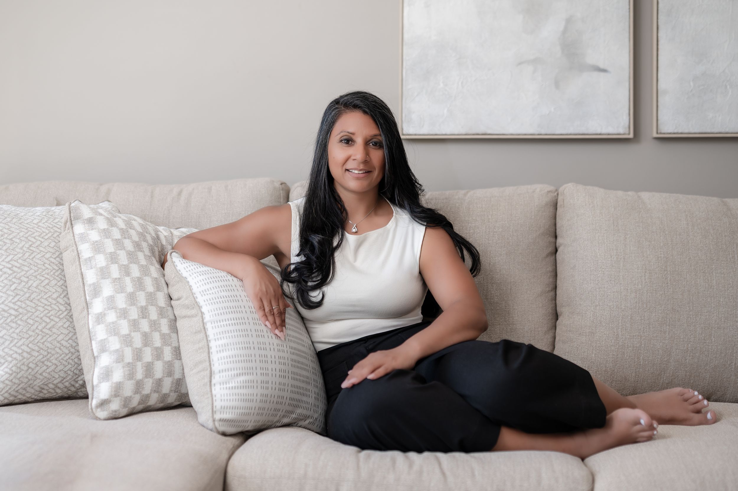 Woman in white top and black pants sitting relaxed on a beige sofa with decorative pillows in a modern living room.