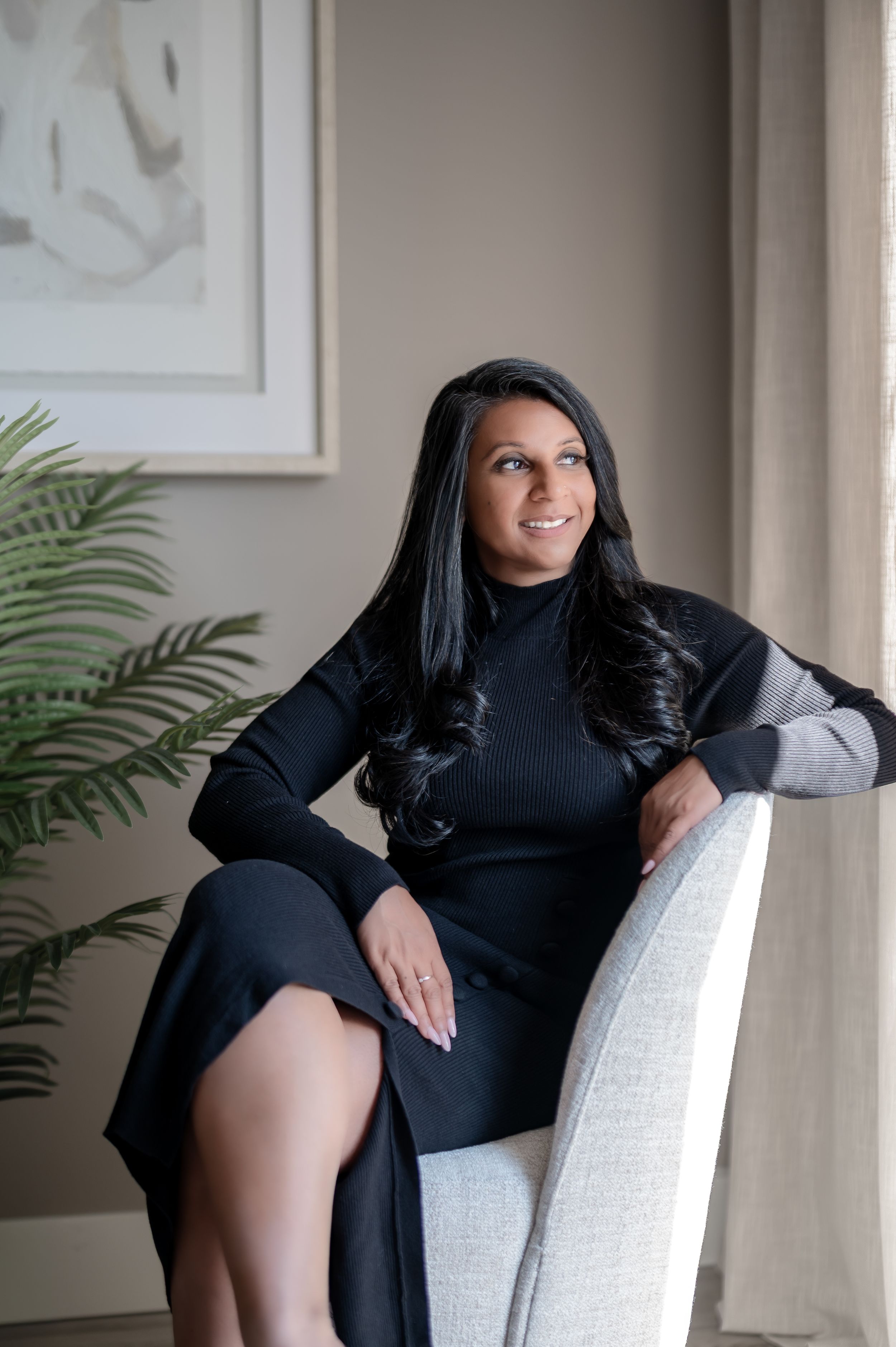 Woman in black turtleneck dress seated elegantly in a white chair, smiling in a modern, softly lit interior.