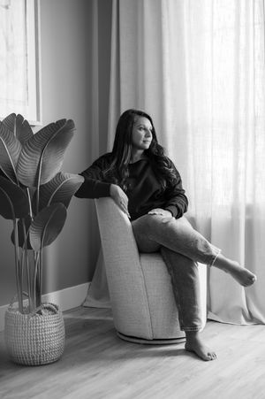 Black and white photo of a woman sitting in a chair by a window with a large plant nearby.