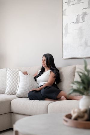 Woman with long dark hair sits relaxed on a beige sofa with decorative pillows in a modern, neutral-toned living room.