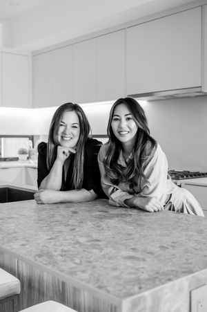 Two smiling women leaning on a marble kitchen island in a modern white kitchen, black and white photo.