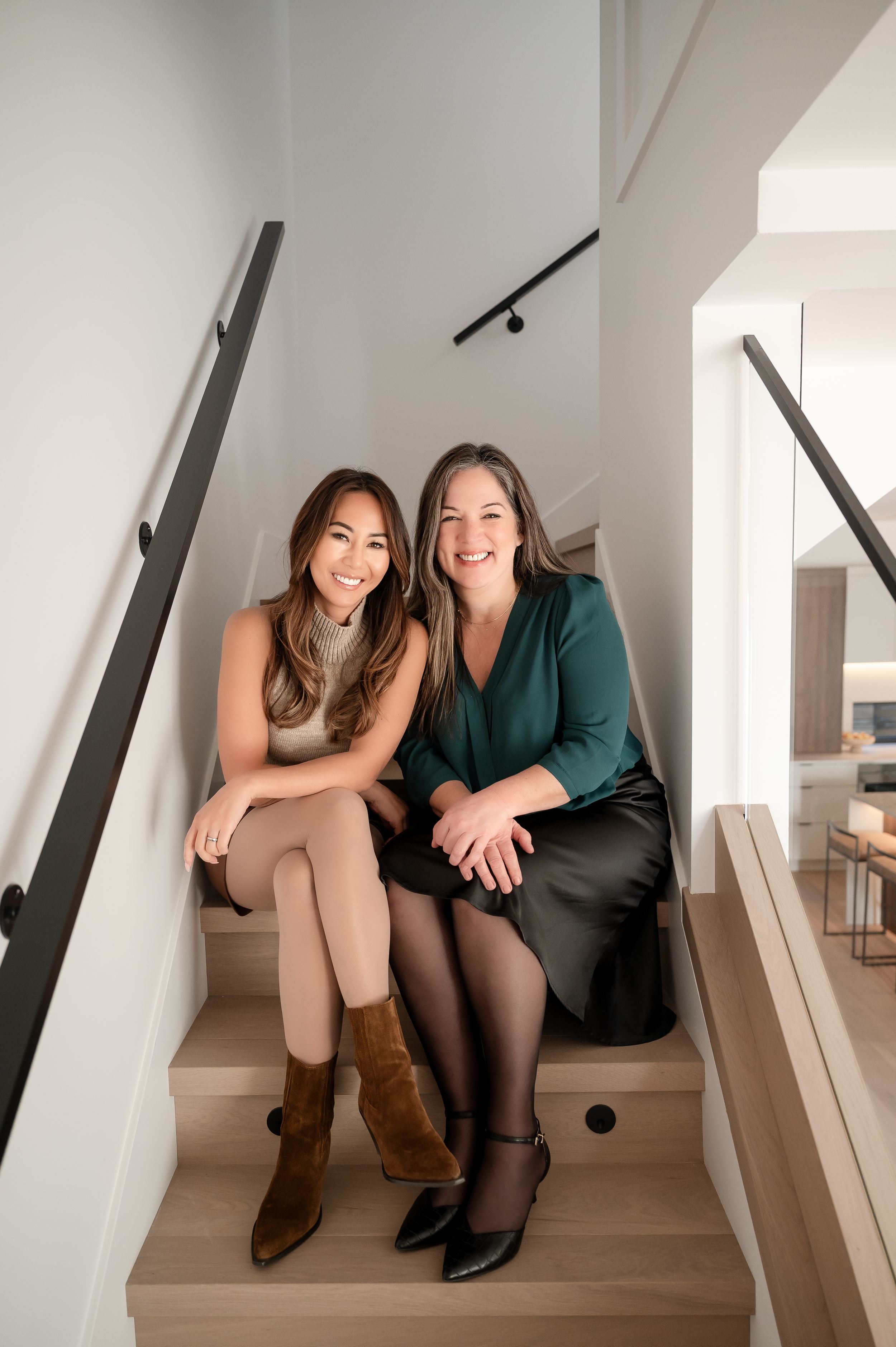 Two smiling women sitting together on modern wooden stairs in a bright contemporary home interior.