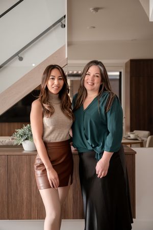 Two stylish women posing together in a modern interior, one in a brown leather skirt and the other in a green blouse.