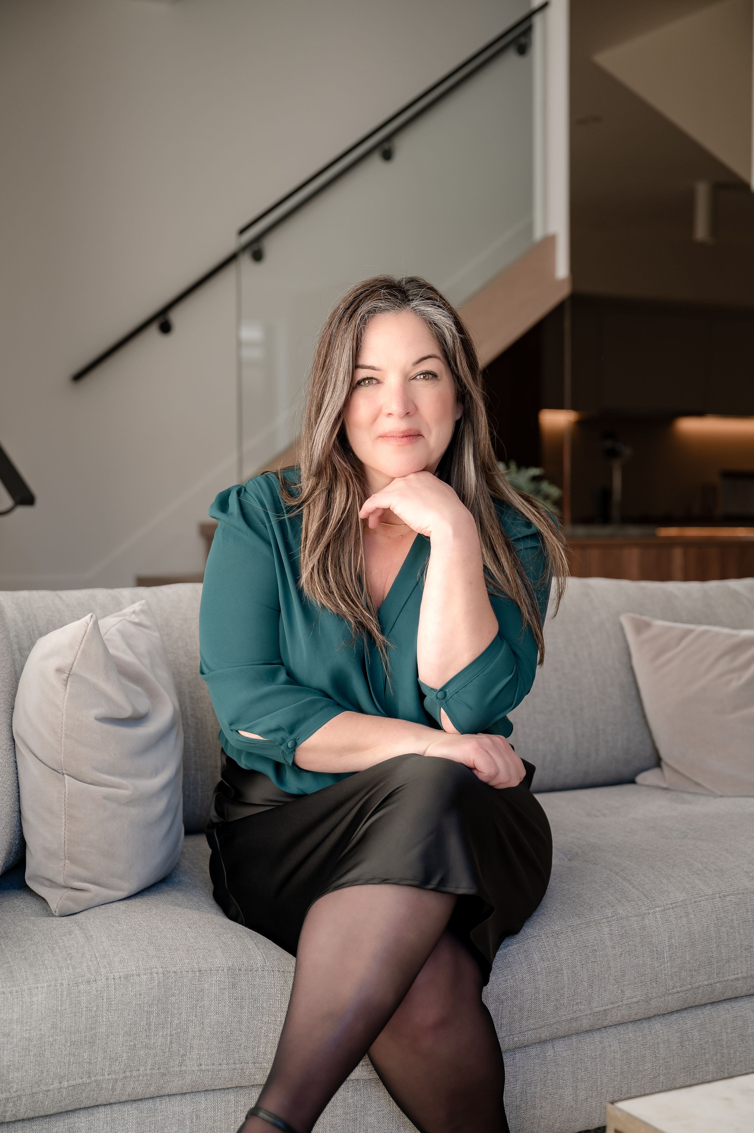 Woman in teal blouse and black skirt sitting on gray sofa in modern home interior with staircase.