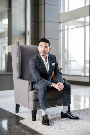 A well-dressed man in a dark plaid suit sits confidently in a gray wingback chair in a modern lobby.