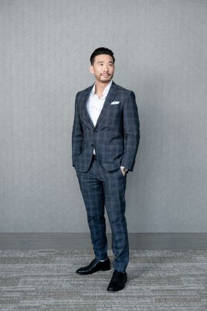 Man in navy plaid suit with white pocket square standing confidently against gray background.