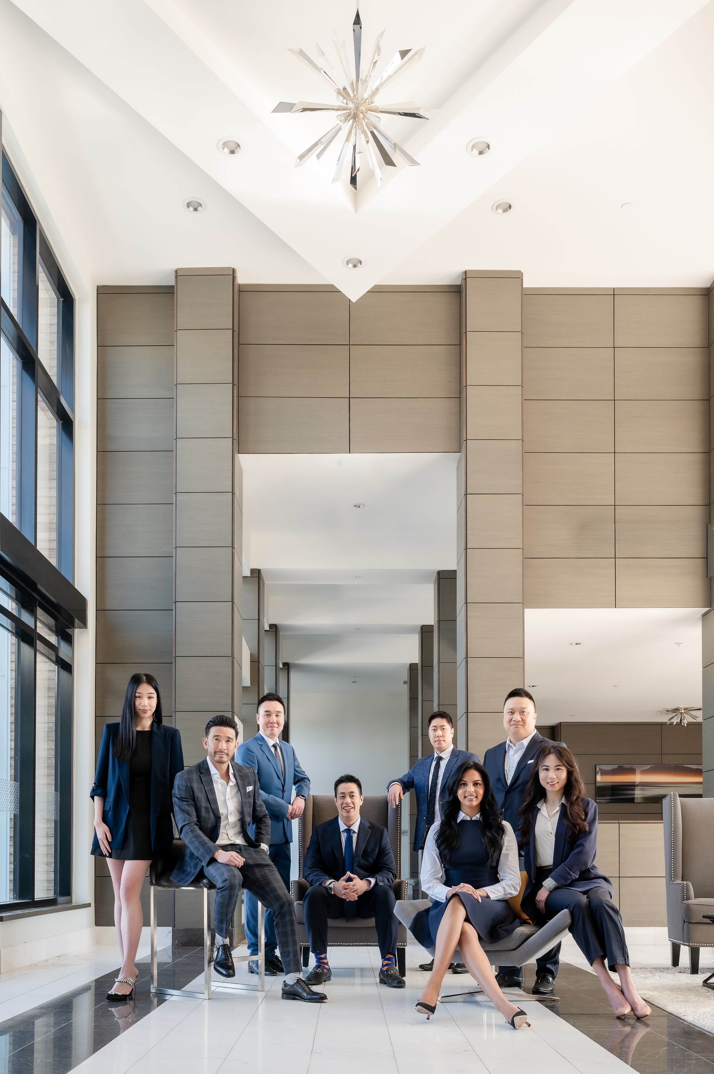 Professional business team of eight posing in a modern corporate lobby with elegant chandelier.