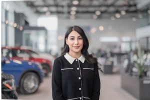 Professional wearing a black cardigan with white trim stands in a modern car dealership showroom with blurred vehicles in background.