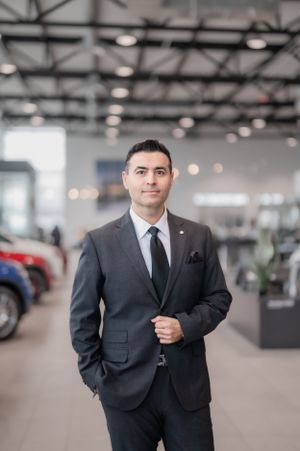 A professional in a dark business suit stands confidently in a modern car dealership showroom with vehicles in background.