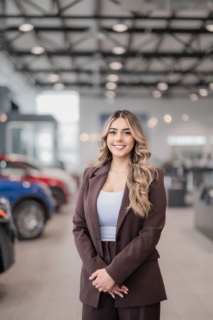 Professional in a brown business suit stands smiling in a car showroom with vehicles and lighting visible in background.
