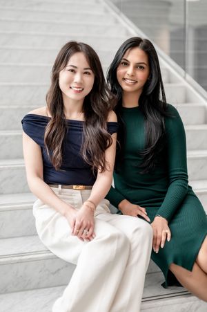 Two women in contrasting navy and green dresses sit together on white stairs in a bright setting.