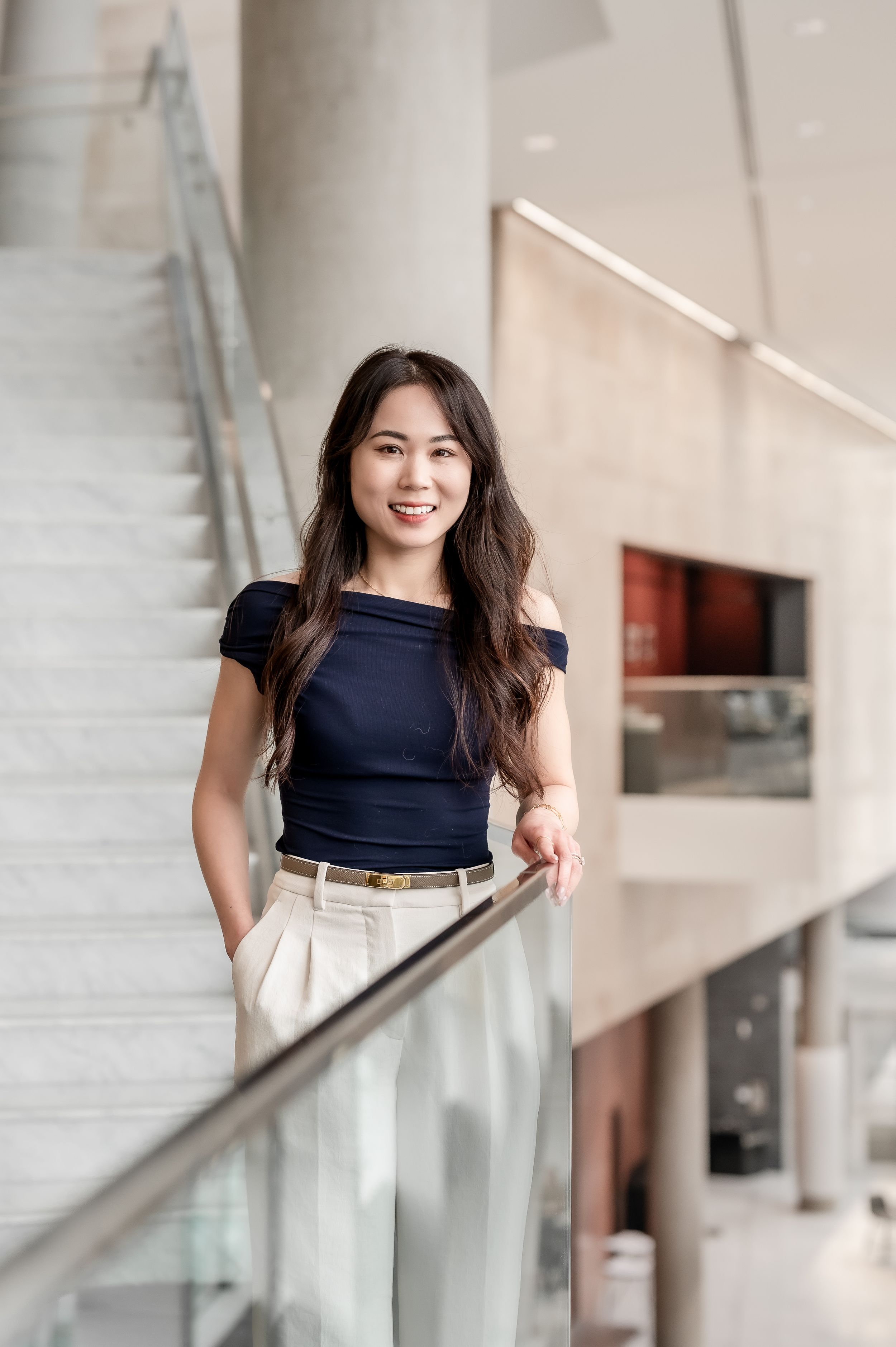 A smiling young woman in a navy off-shoulder top and cream trousers poses by a modern glass railing indoors.