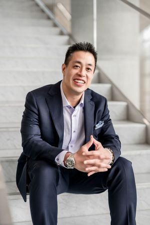 Smiling man in navy suit sitting on marble stairs with hands clasped, wearing a watch and pocket square.