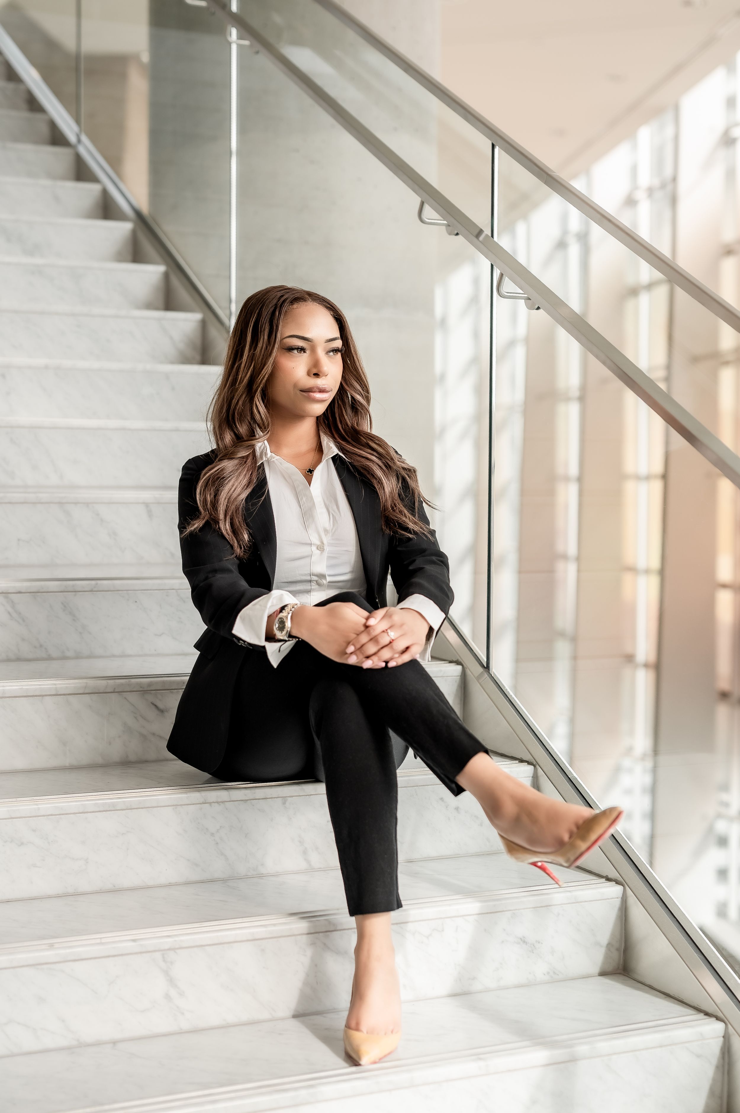 Professional business attire worn with nude heels while sitting elegantly on modern marble stairs with glass railing.