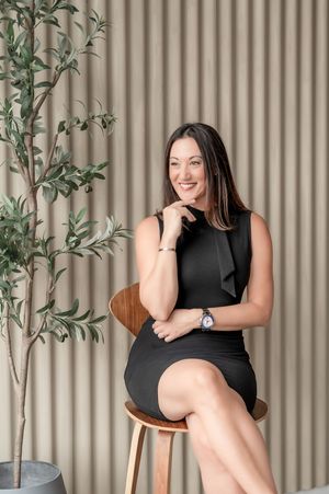 A woman in a black dress poses on a wooden chair against a beige corrugated wall with green plants.