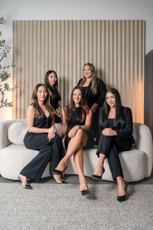 Four women in black outfits pose casually on a modern gray sofa against a beige wall with soft lighting.