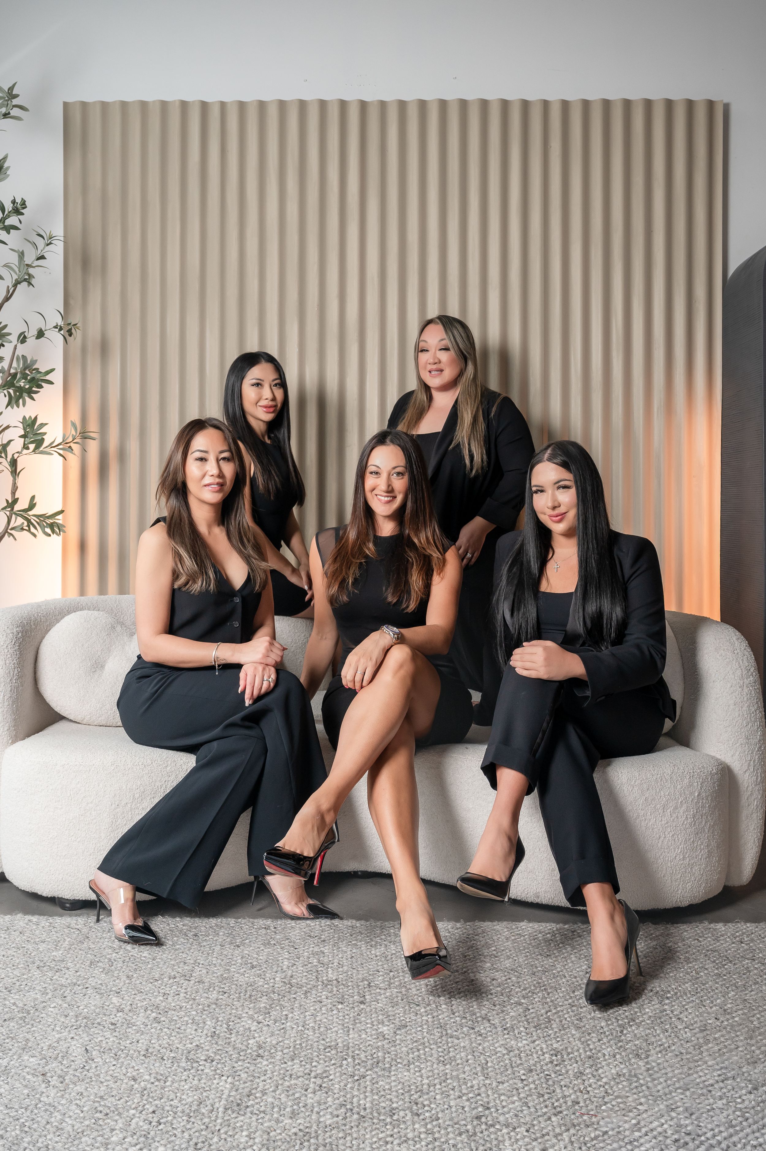 Four women in black outfits pose casually on a modern gray sofa against a beige wall with soft lighting.