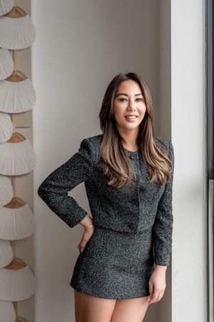 Professional portrait of a woman in a dark tweed blazer dress standing by a window with decorative wall hangings.