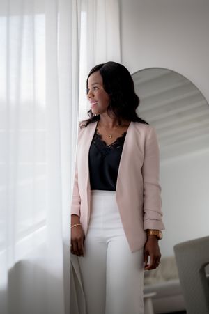 Professional woman wearing a soft pink blazer and white pants stands near curtained window in bright modern space.