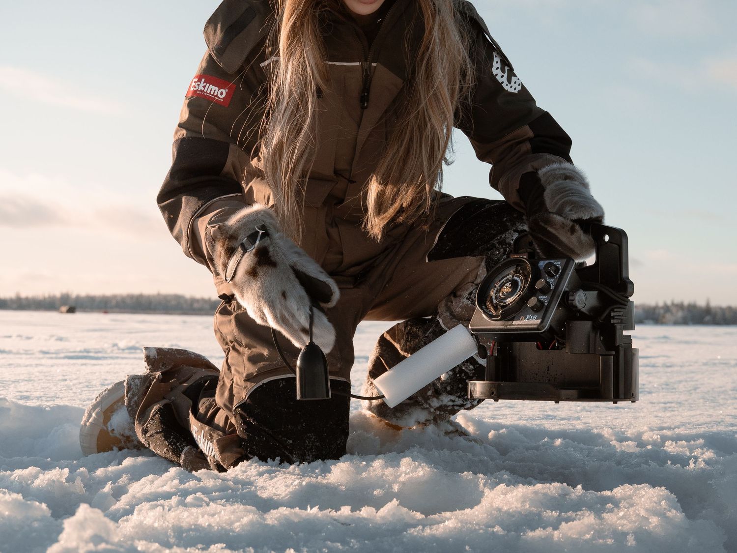 Person kneeling in snow while operating ice fishing equipment on frozen lake surface.